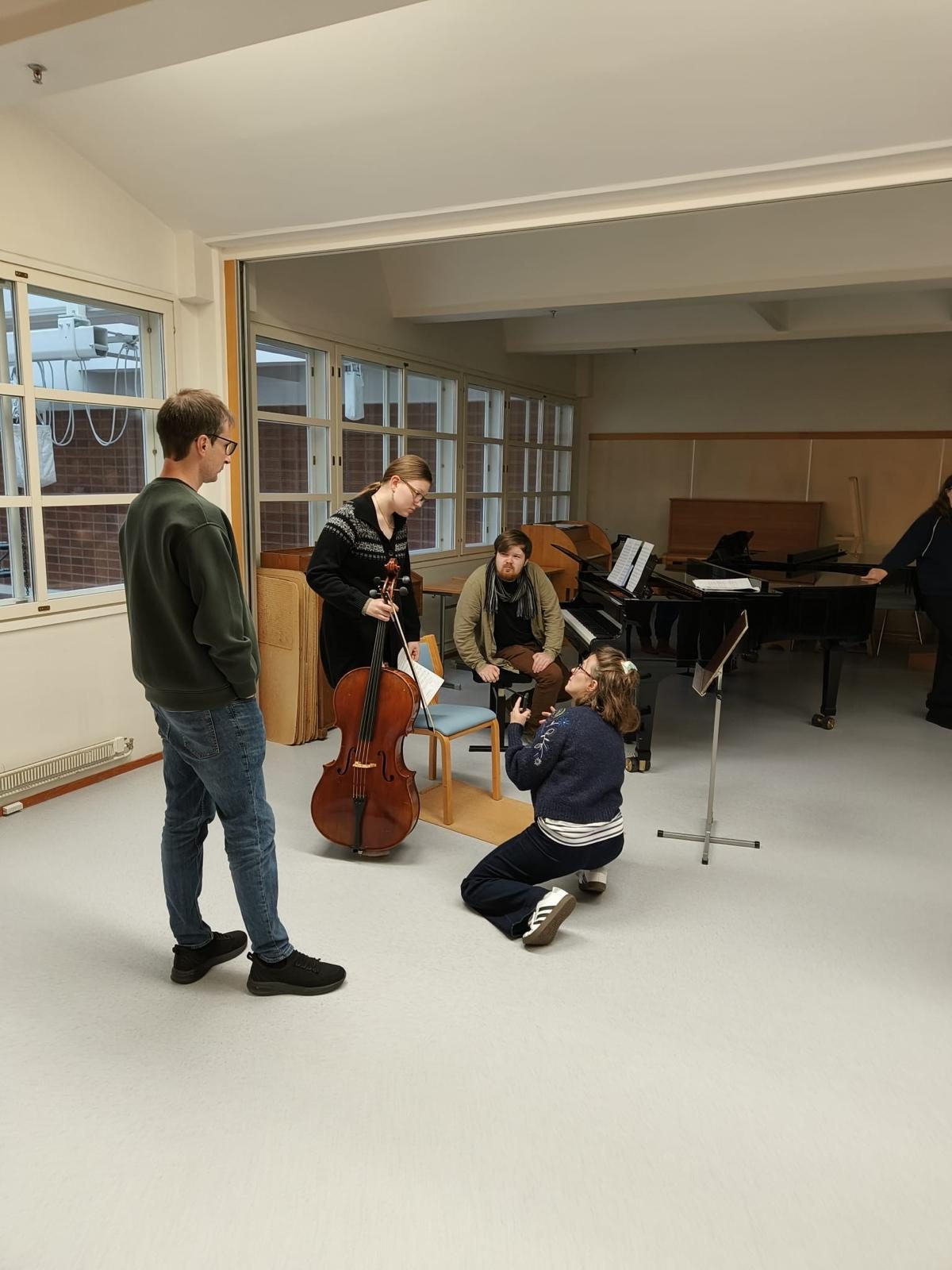 Josefina Urraca kneeling to work with a cellist while Guillermo Laporta watches, in a conservatory rehearsal room