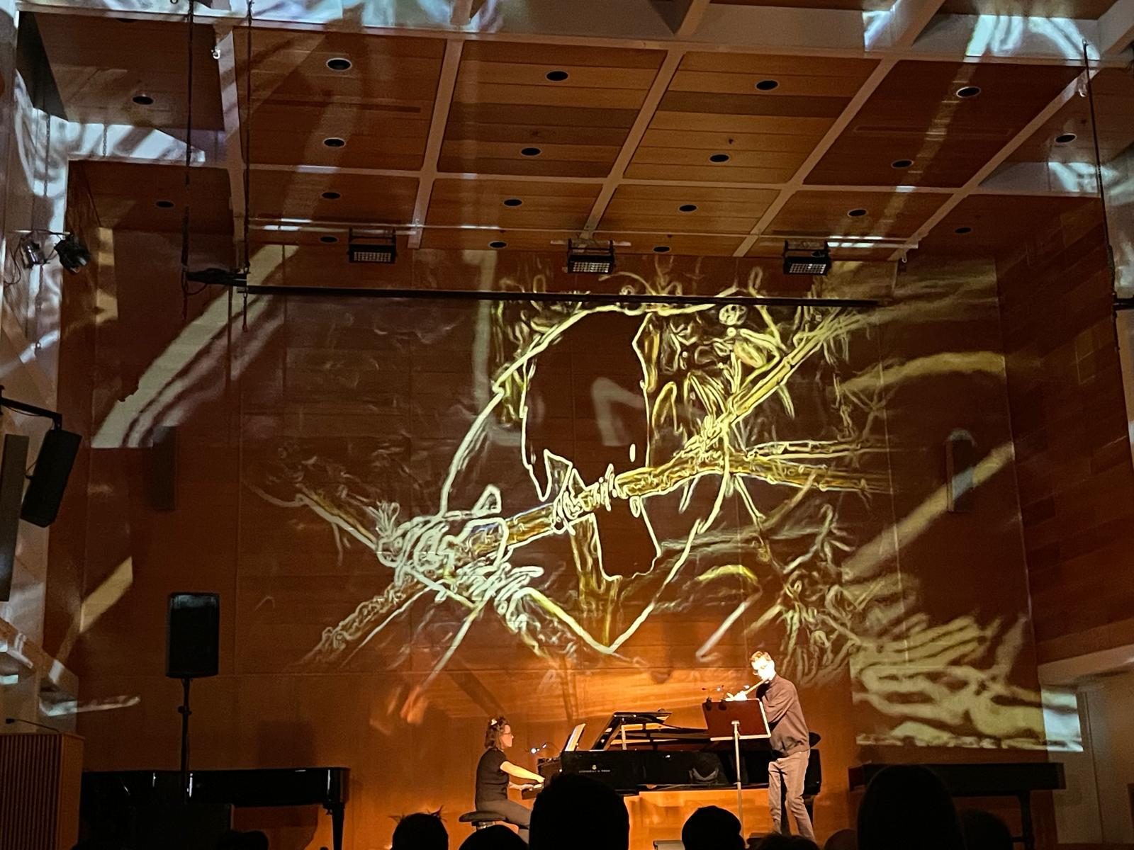 Josefina Urraca at the piano and Guillermo Laporta on flute, with a projection filling the wall of the hall